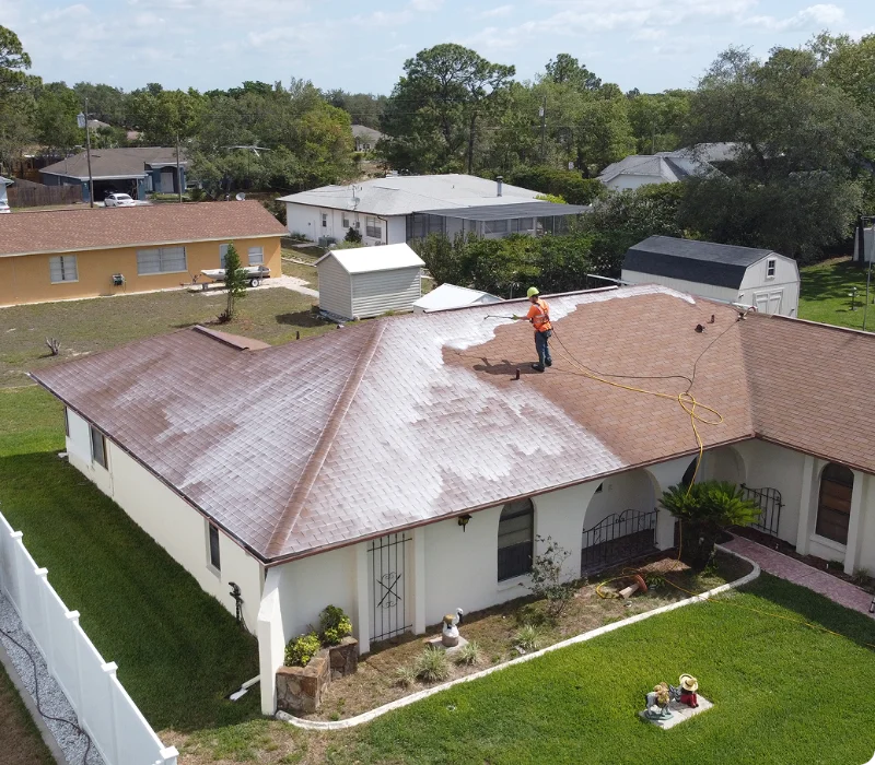 A person in an orange shirt sprays cleaning solution onto a one-story roof using a long yellow hose, creating white foam. The roof rejuvenation is part of an STL Roof Renew project in a suburban St Louis neighborhood with green lawns and trees.