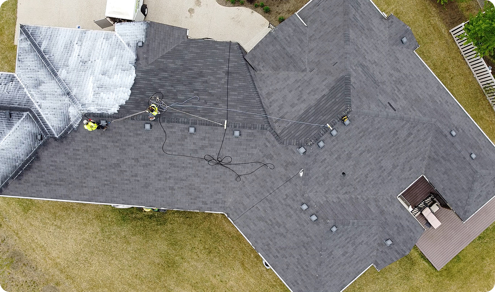 Aerial view of STL Roof Renew workers in St Louis cleaning or repairing the dark gray shingled roof of a large house. Hoses and equipment are spread across the roof, with green grass and a small paved area surrounding the property.