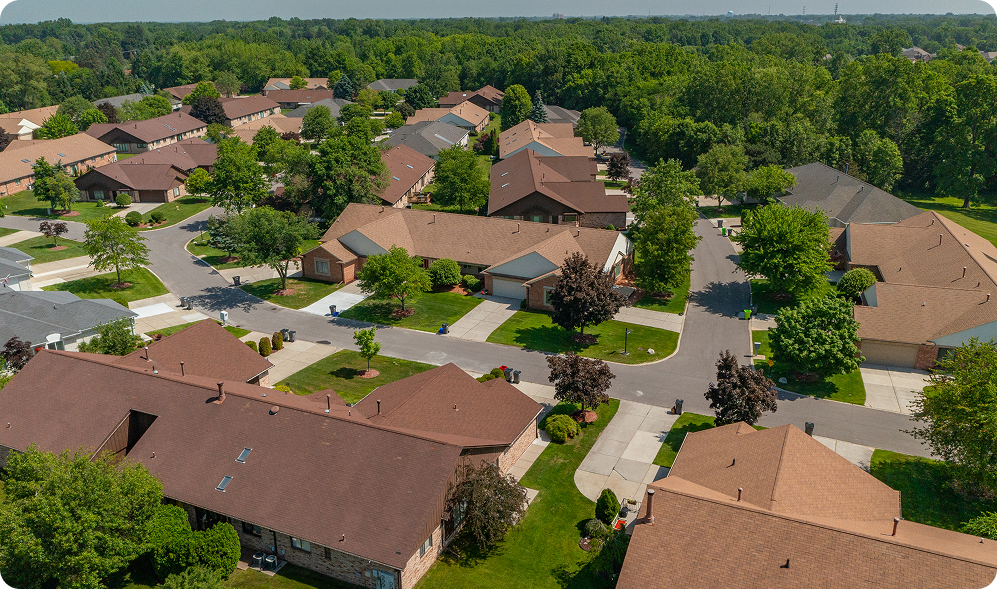 Aerial view of a suburban St Louis neighborhood with rows of single-family houses, brown roofs, green lawns, trees lining the streets, and a background of dense forest. The sky is clear and sunny—perfect for STL Roof Renew and roof rejuvenation.