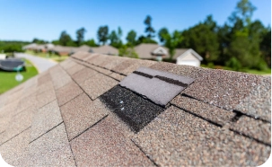 Close-up of a residential roof in St Louis with missing or damaged shingles, exposing black underlayment beneath. Trees, houses, and a clear blue sky form the blurred background—ideal for a STL Roof Renew or Roof Rejuvenation service.